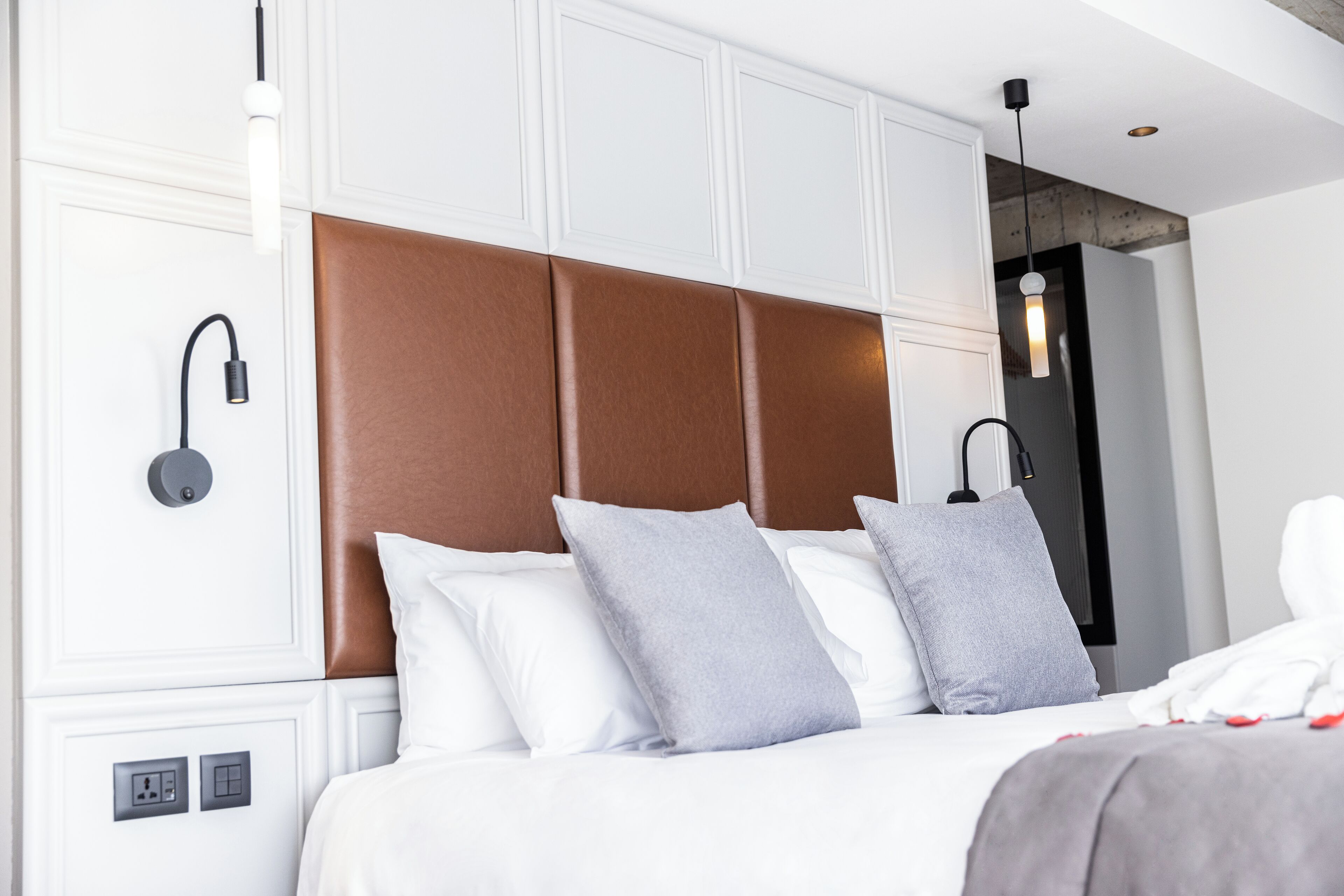 A modern hotel room with a large bed, white walls, and a brown headboard. There are two bedside tables with lamps.