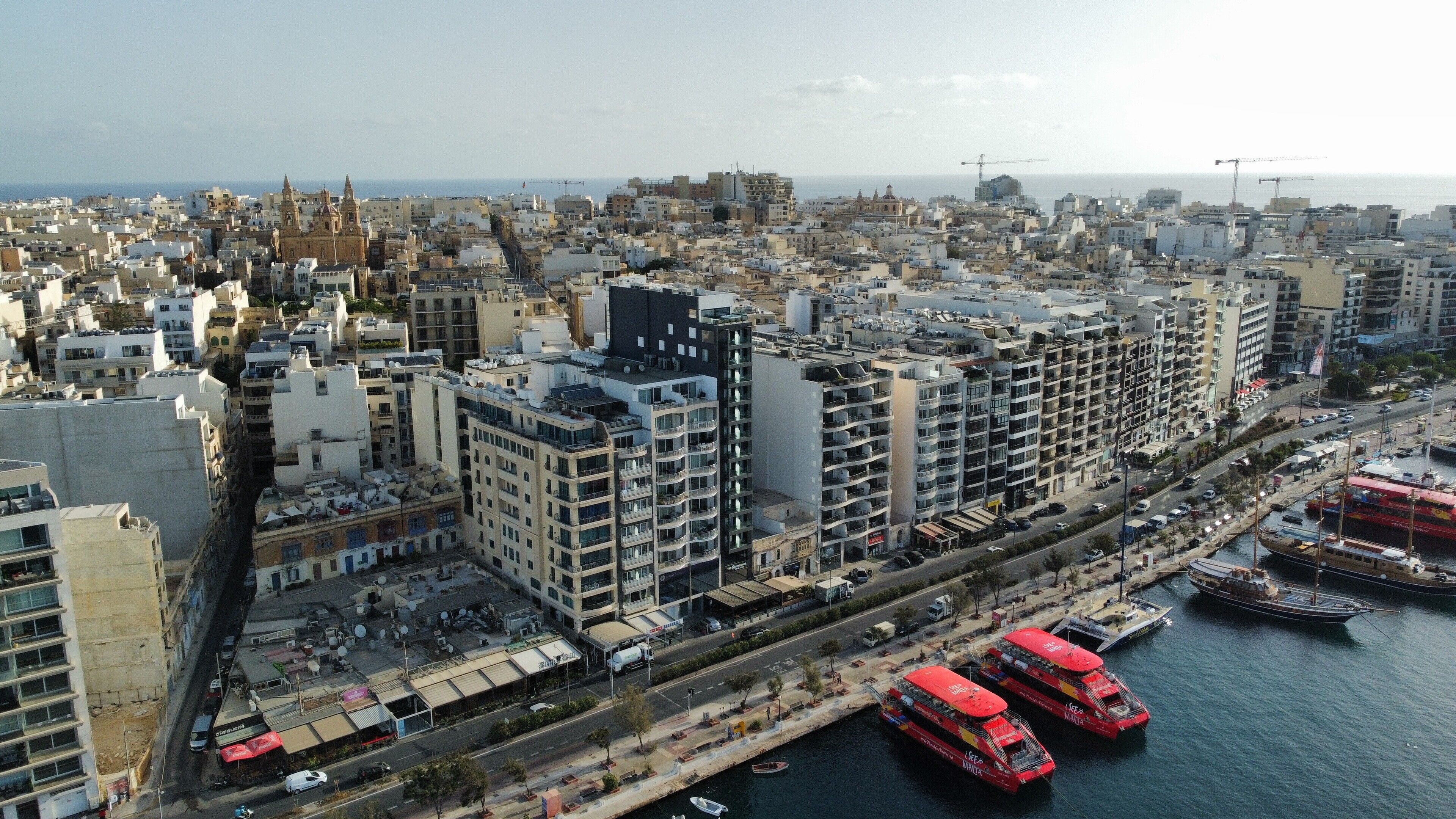 Un paisaje urbano costero con edificios densos, una iglesia destacada y un puerto deportivo con barcos rojos.