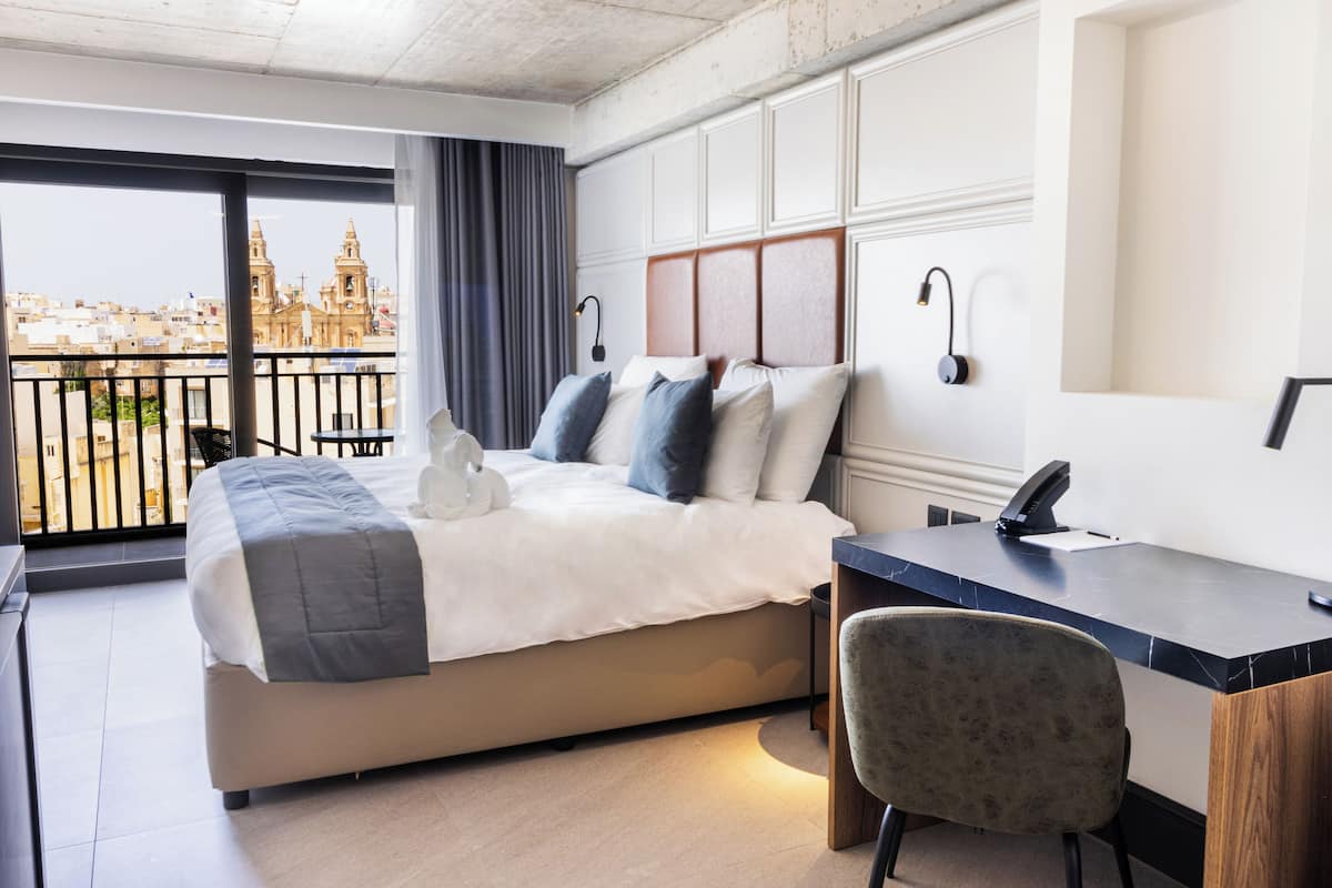 Habitación de hotel con cama, escritorio, silla y vistas a un paisaje urbano a través de amplios ventanales.