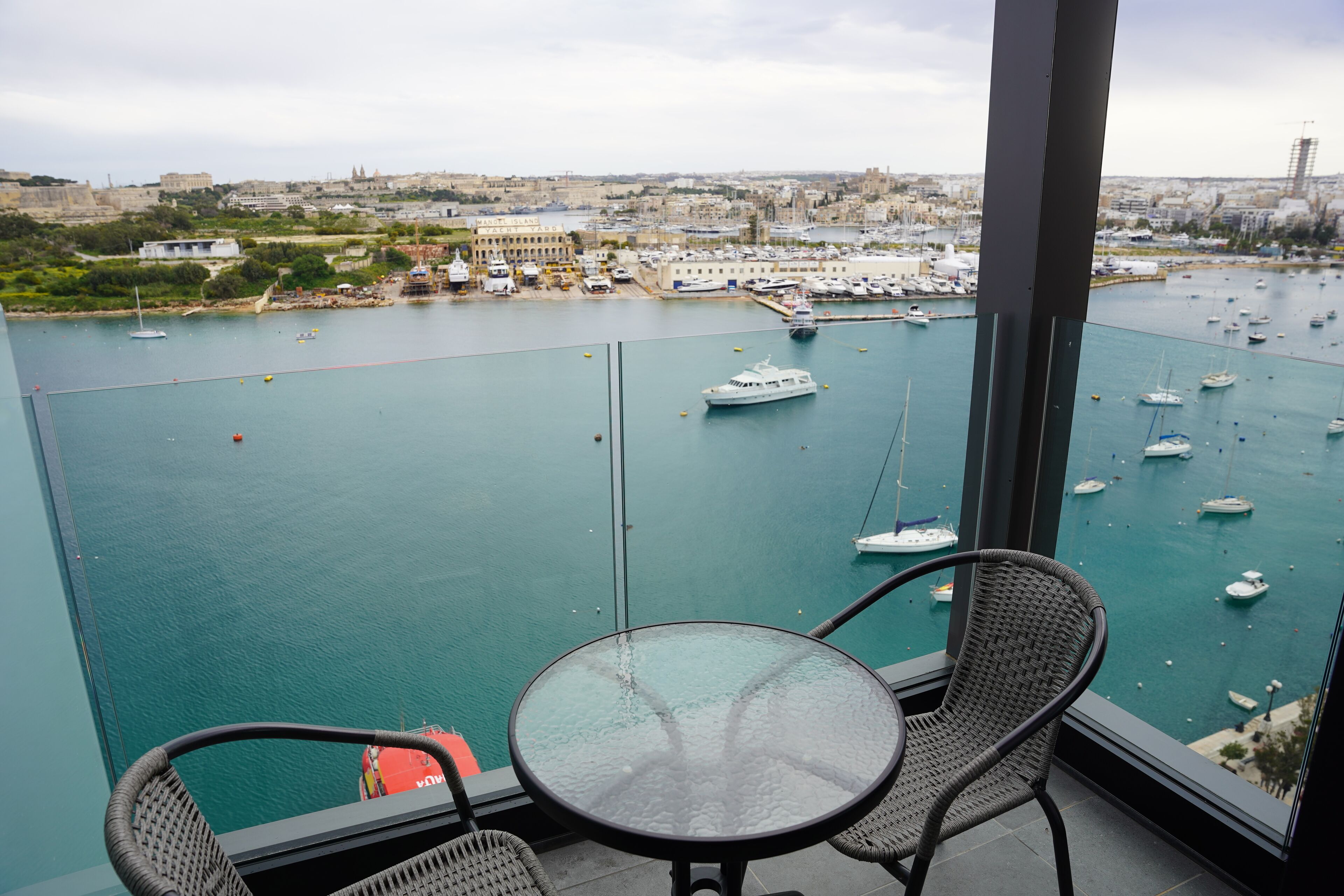 A balcony with a glass table and wicker chairs overlooking a marina with boats.