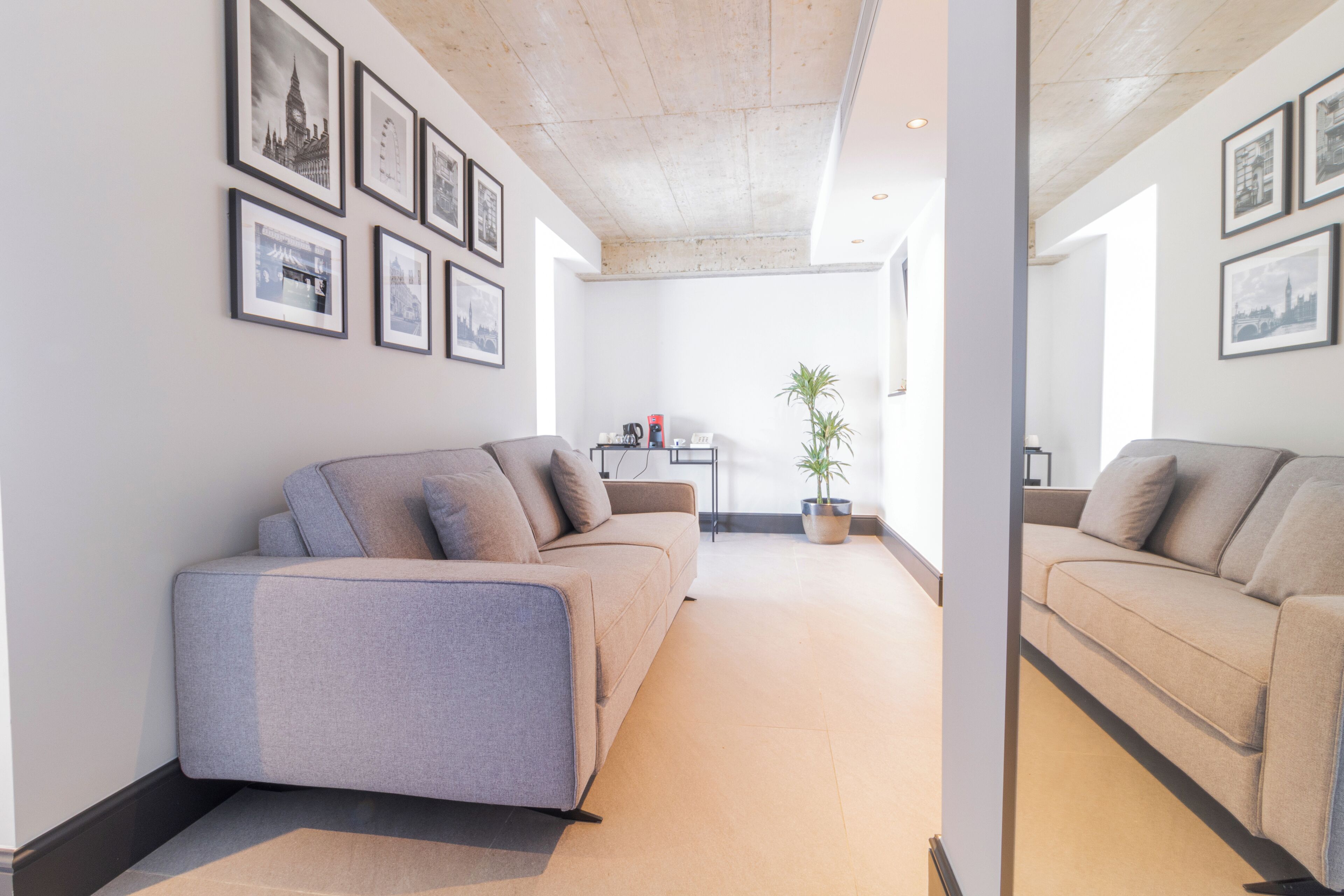 A modern living room with a grey sofa, a coffee table, and a potted plant. There are framed pictures on the wall and a window providing natural light.