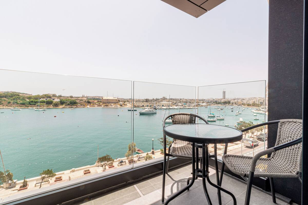 Un balcón con vista a un puerto deportivo, que incluye barcos y un paisaje urbano distante.