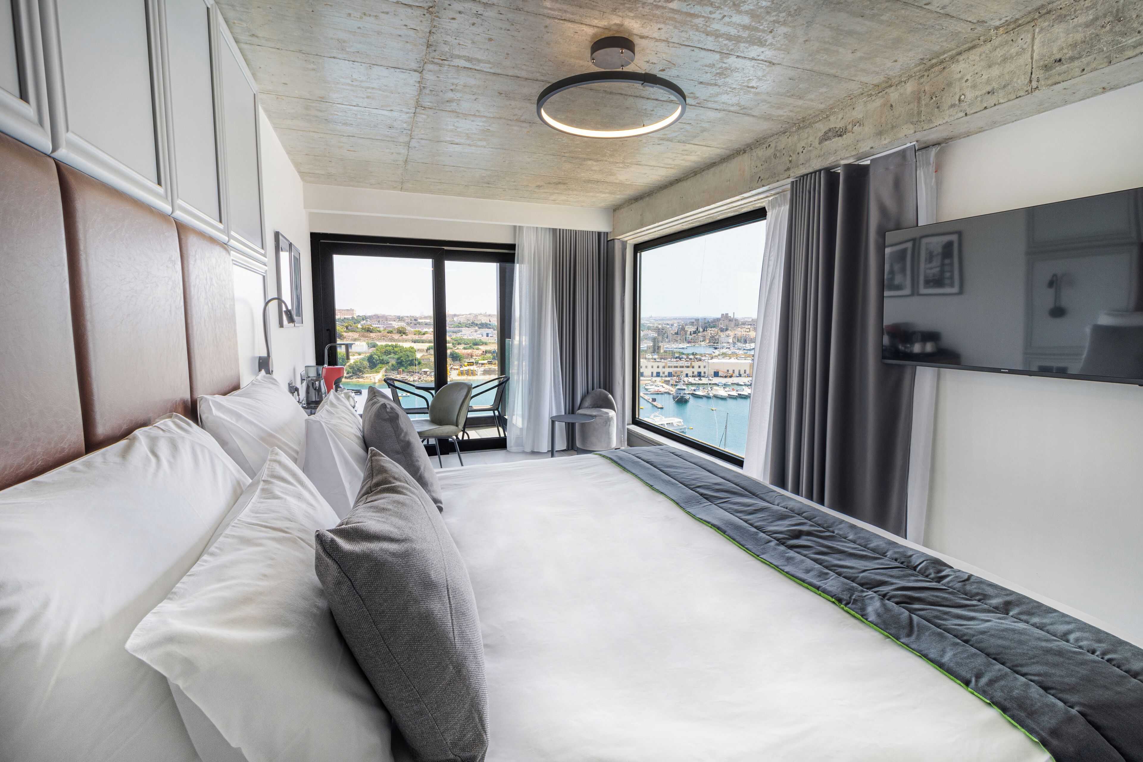 A modern hotel room with a large bed, a view of the city, and a balcony with a view of the harbor.