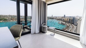Une chambre d’hôtel moderne dotée d’un grand balcon, d’un coin repas et offrant une vue sur un port de plaisance et le paysage urbain.