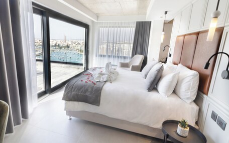 A modern hotel room with a large bed, a chair, a small table with a plant, and a view of the city through a large window.