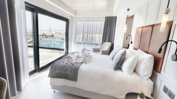 Une chambre d’hôtel moderne avec un grand lit, une chaise, une petite table avec une plante et une vue sur la ville, visible à travers une grande fenêtre.