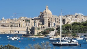 Un paysage urbain côtier avec un bâtiment historique remarquable, surmonté d’un dôme, entouré d’immeubles résidentiels et d’un port de plaisance où sont amarrés différents types de bateaux.