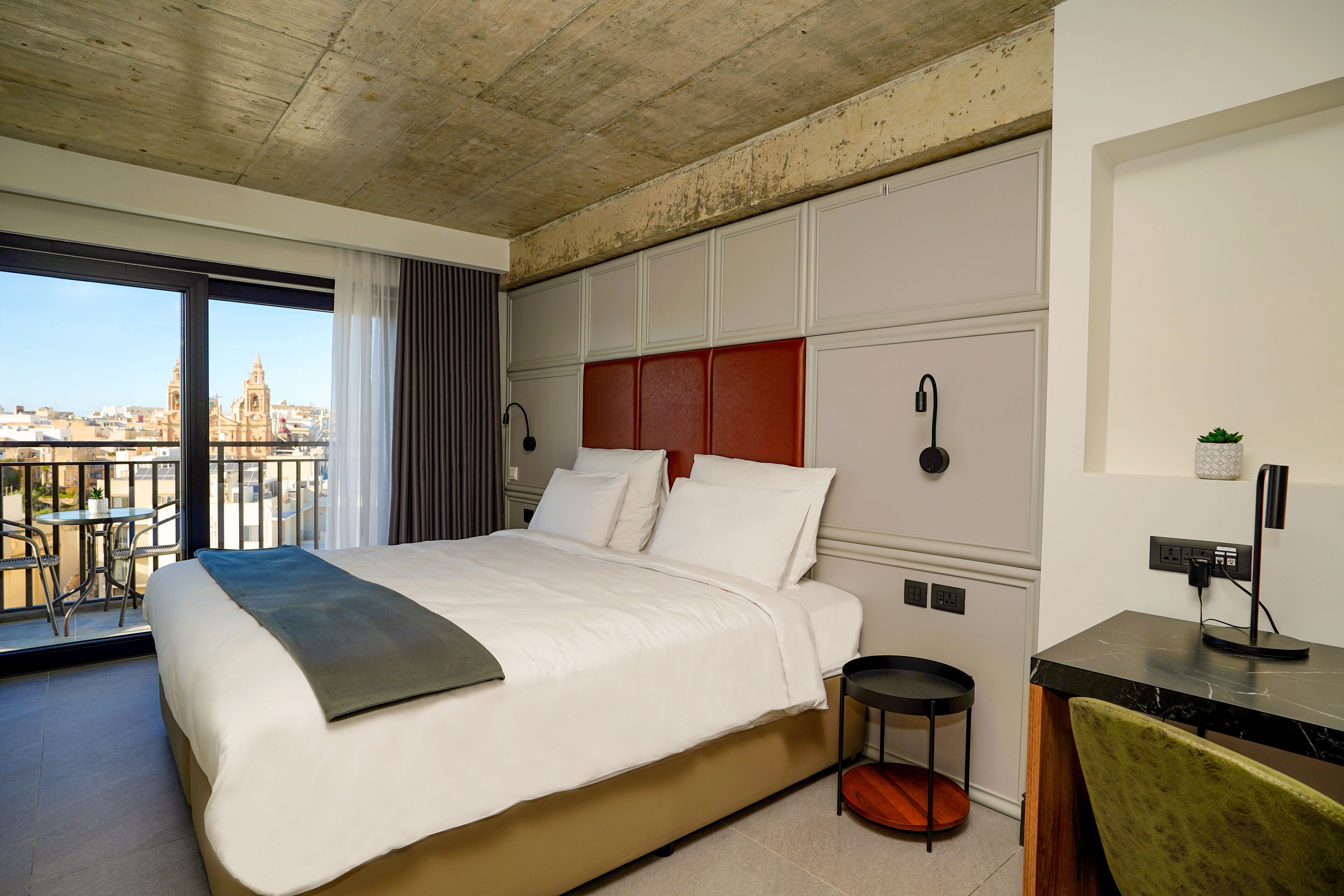 A hotel room with a large bed, a small round table, a desk with a computer, and a view of a cityscape through a balcony.