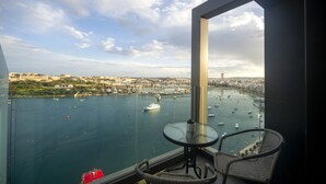 Un balcon avec une table et des chaises en verre, donnant sur un port où sont amarrés des bateaux et sur une vue panoramique de la ville.
