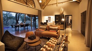 A spacious living area with a large window overlooking a deck, a cozy seating area with a sofa and armchairs, and a kitchen in the background. - Serondella Safari Lodge (Hoedspruit)