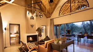 A modern living room with a fireplace, wooden furniture, and large windows overlooking a forest. - Serondella Safari Lodge (Hoedspruit)