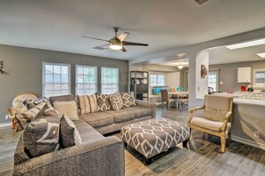 A spacious living room with a gray sectional sofa, ottoman, and a wooden chair. A ceiling fan and large windows provide natural light. - Copy 3 Converse Home-12 Mi to San Antonio River Walk (Converse)