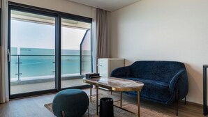 A modern room with a large sliding glass door leading to a balcony with a view of the sea. There is a blue sofa, a small round table, and a white minibar. - HOTEL GITE (Siheung)