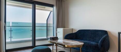 Una habitación moderna con un gran ventanal corredero que da a un balcón con vistas al mar. Hay un sofá azul, una mesita redonda y un minibar blanco.