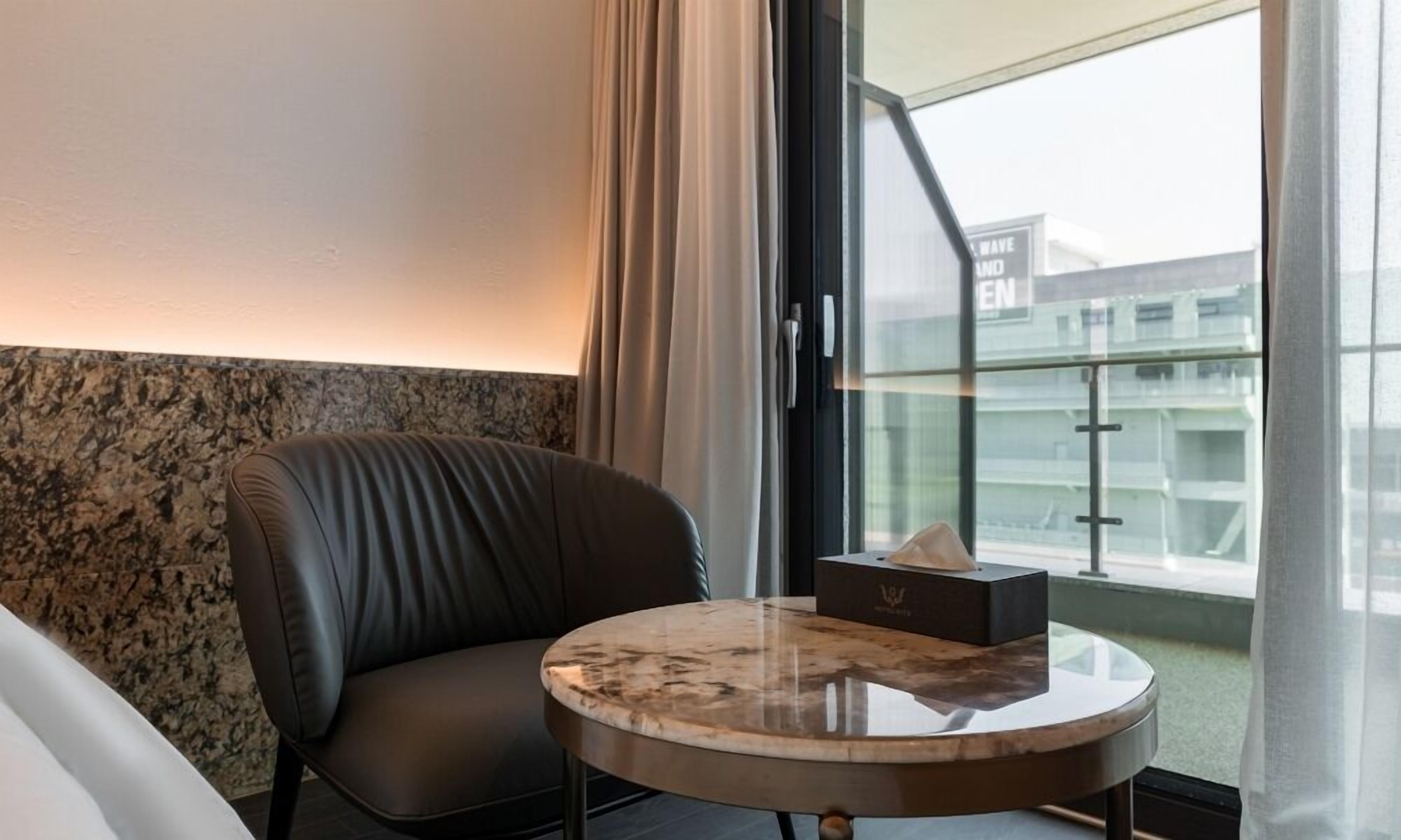 Camera d'albergo con una poltrona in pelle, un tavolino rotondo in marmo e vista su un edificio attraverso un'ampia finestra.