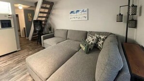 Un salon moderne avec un canapé gris, un escalier menant à une autre pièce et un coin cuisine visible au fond.