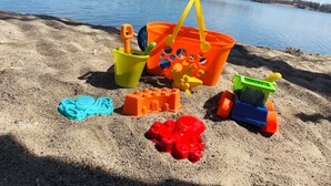 En strandscene med sandlegetøj, en gul spand og en blå vandmasse i baggrunden.