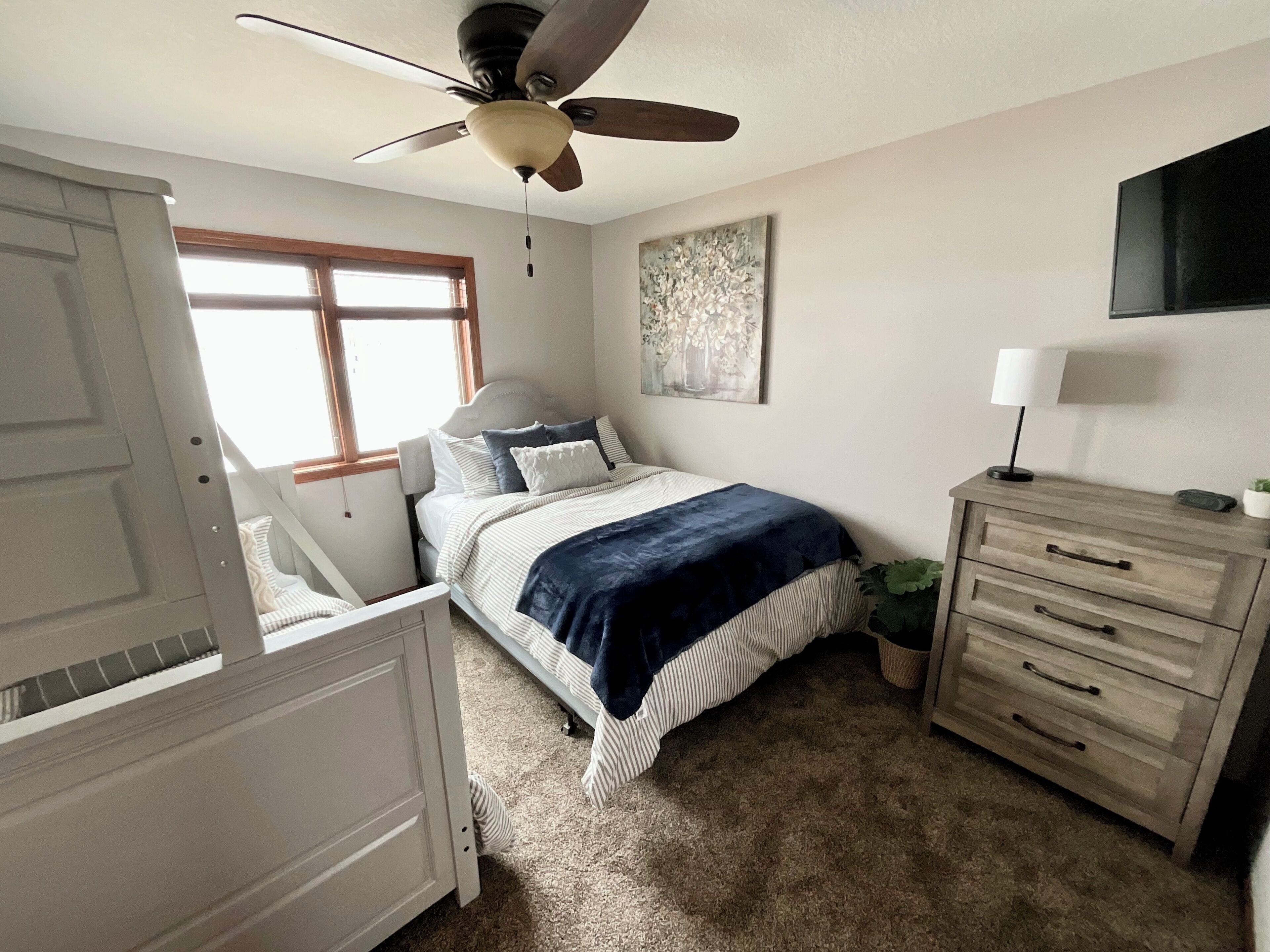 A bedroom with a bed, a dresser, a ceiling fan, a window, and a painting on the wall.