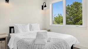 A neatly made bed with white bedding and a folded towel, a nightstand with a lamp, and a window with a view of trees. - Long Shadow - MTB Oasis (Bella Vista)