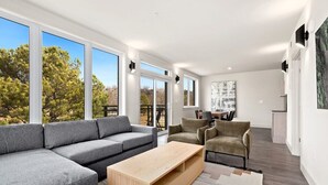 Ein modernes Wohnzimmer mit einem grauen Ecksofa, zwei Sesseln, einem Holztisch und einem großen Fenster mit Blick auf Bäume.