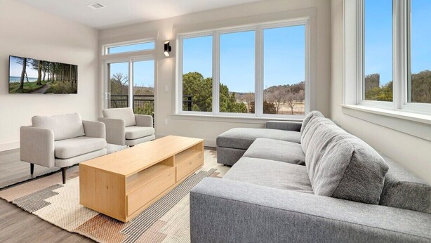 Een moderne woonkamer met een grijze modulaire bank, twee fauteuils, een houten salontafel en een groot raam met uitzicht op bomen en heuvels.
