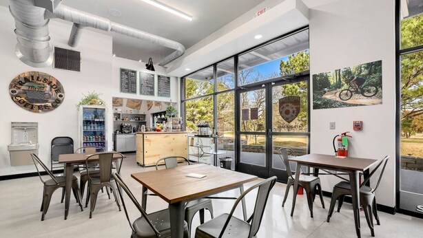 Interior moderno de café com mesas, cadeiras, um quadro de cardápio e grandes janelas com vista para o exterior.
