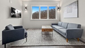 Un salon moderne avec un canapé gris, une table basse et deux fauteuils. Une grande fenêtre offre une vue sur les arbres, et un tableau est accroché au mur.