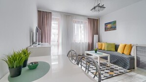 Een moderne woonkamer met een bank, een eettafel en stoelen. Er staat een tv op een dressoir, een schilderij aan de muur, en een raam met gordijnen.