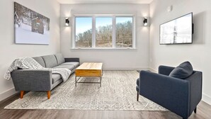 Un salon moderne avec un canapé gris, un fauteuil bleu, une table basse en bois et une grande fenêtre donnant sur des arbres.