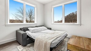 Ein Schlafzimmer mit einem Bett, einem grauen Sofa, einem Holztisch und großen Fenstern mit Blick auf Bäume und Gebäude.