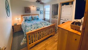 Une chambre avec un lit en bois, une table de chevet, un ventilateur de plafond et un lave-linge dans un coin.