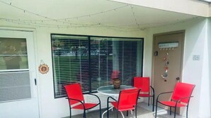 Un patio couvert comprenant une table ronde et quatre chaises rouges, de grandes fenêtres équipées de stores et une porte agrémentée d’un élément décoratif.