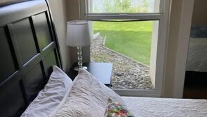 A neatly made bed with white linens and a colorful pillow, a bedside lamp, and a view of a green landscape through a window. - #181 Condo near large pool & shuttle to Gorge Amp-book separately (Quincy)