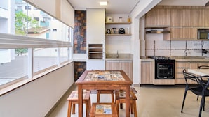 A modern kitchen with a wooden table and stools, a built-in oven, and a view of the outside. - Edífico Residencial Unique (Ubatuba)