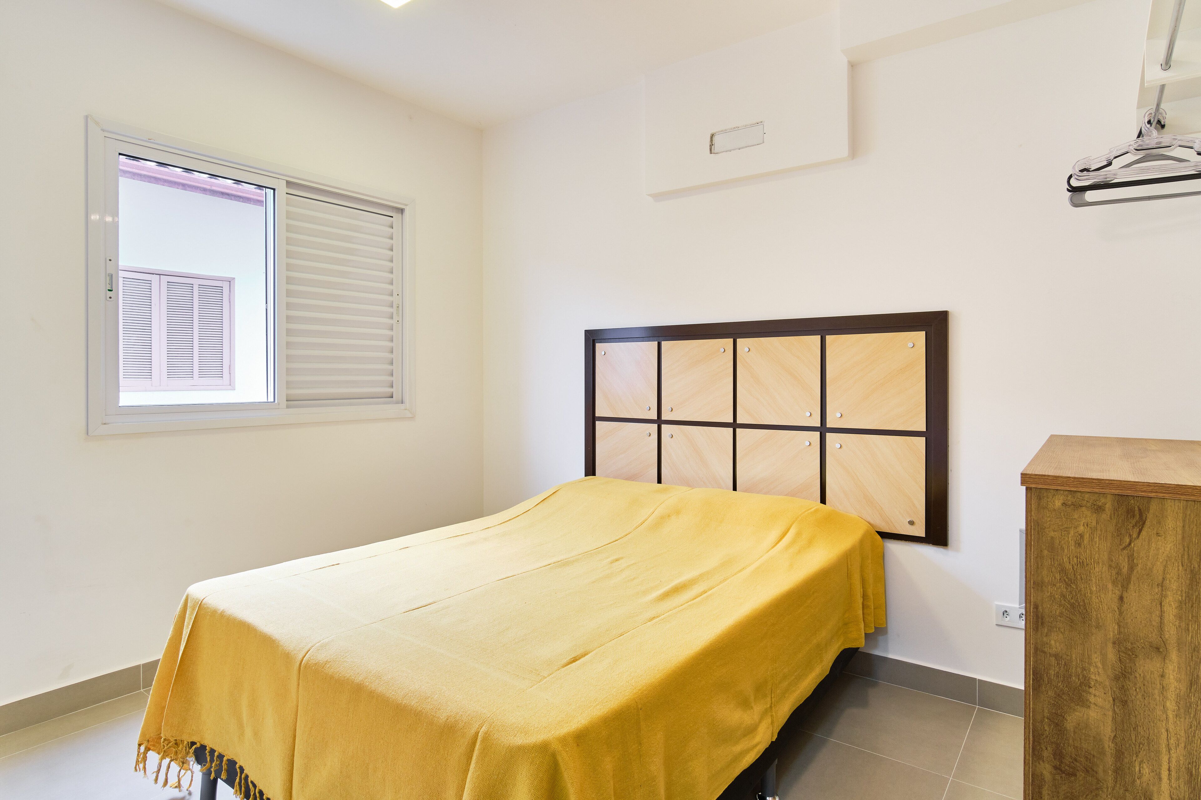 A bedroom with a bed, a window with blinds, a wooden headboard, and a wooden cabinet.