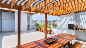 A wooden pergola with a table and bench, potted plants, and a view of buildings in the background. - Edífico Residencial Unique (Ubatuba)