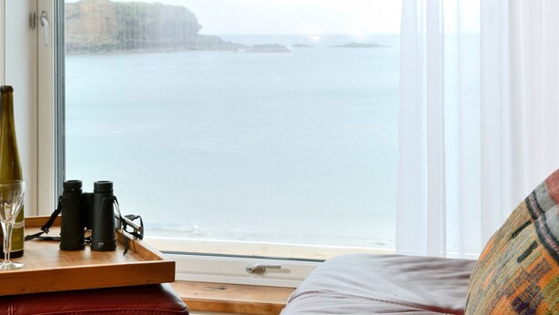 A room with a large window overlooking a coastal view, a wooden tray with binoculars and a wine glass, and a patterned pillow. - Generals Yard (Eyemouth)