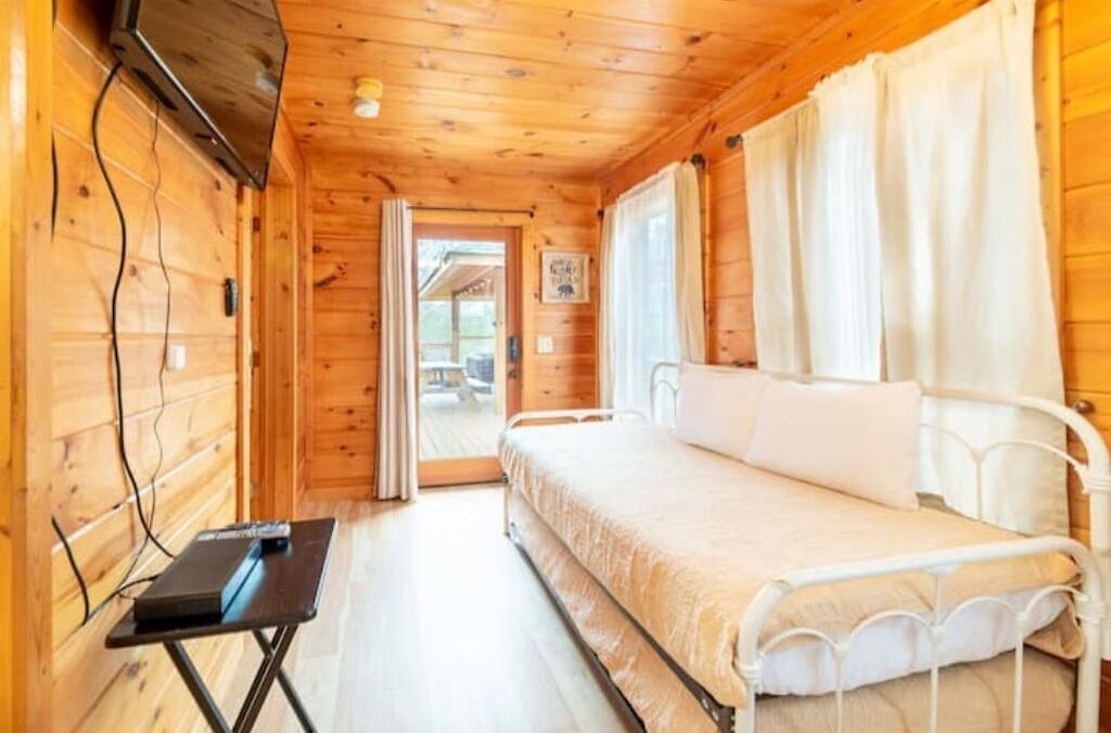 A wooden cabin room with a bed, a small table, and a window with curtains.