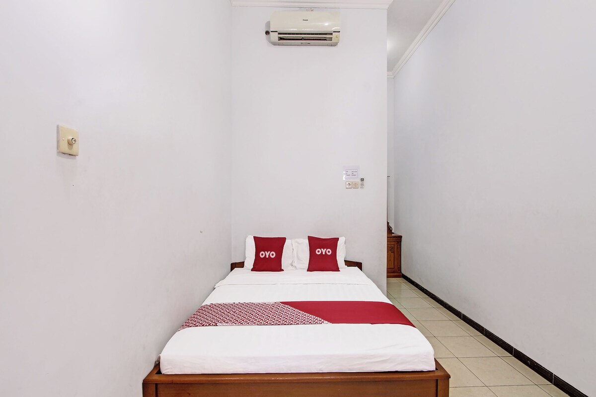 Ein schmales Hotelzimmer mit Bett, weißen Wänden, Holzboden und Klimaanlage über dem Bett.