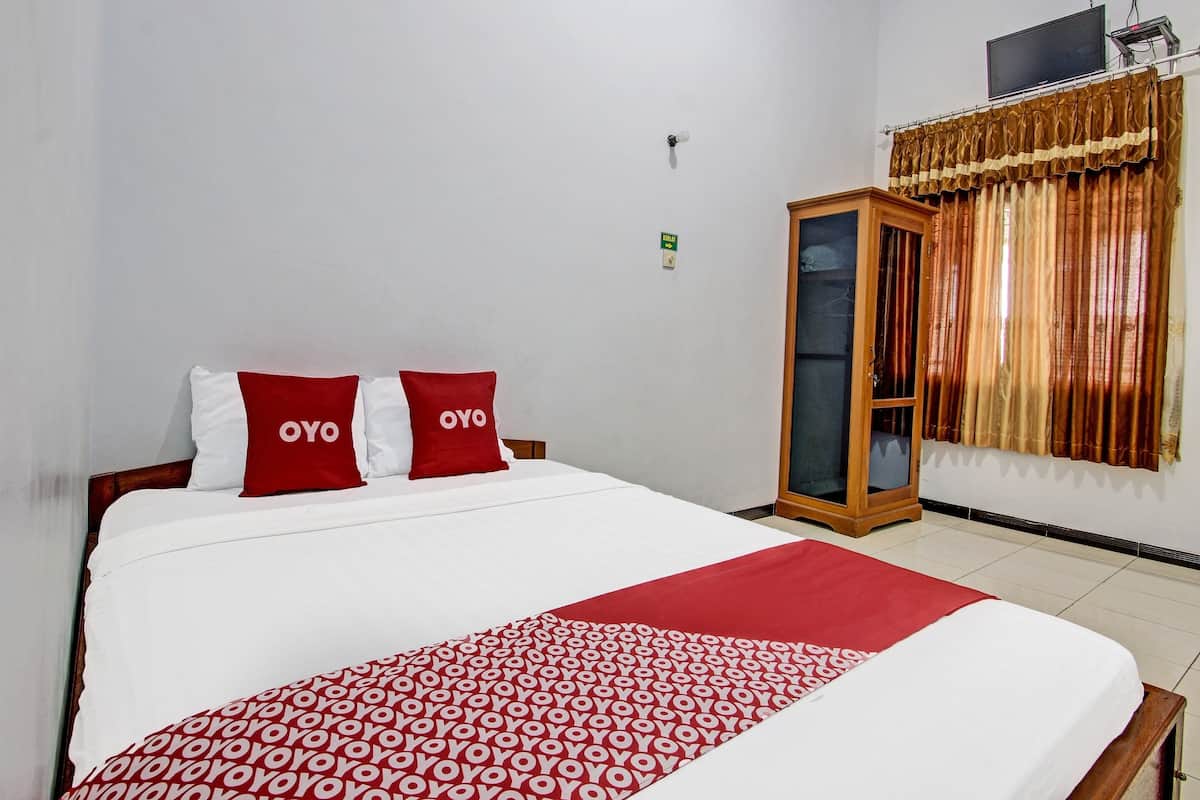 Une chambre d’hôtel avec un grand lit, deux oreillers rouges ornés du mot « OYO », une couvre-lit rouge et blanche à motifs, une armoire en bois et un rideau à motifs dorés.