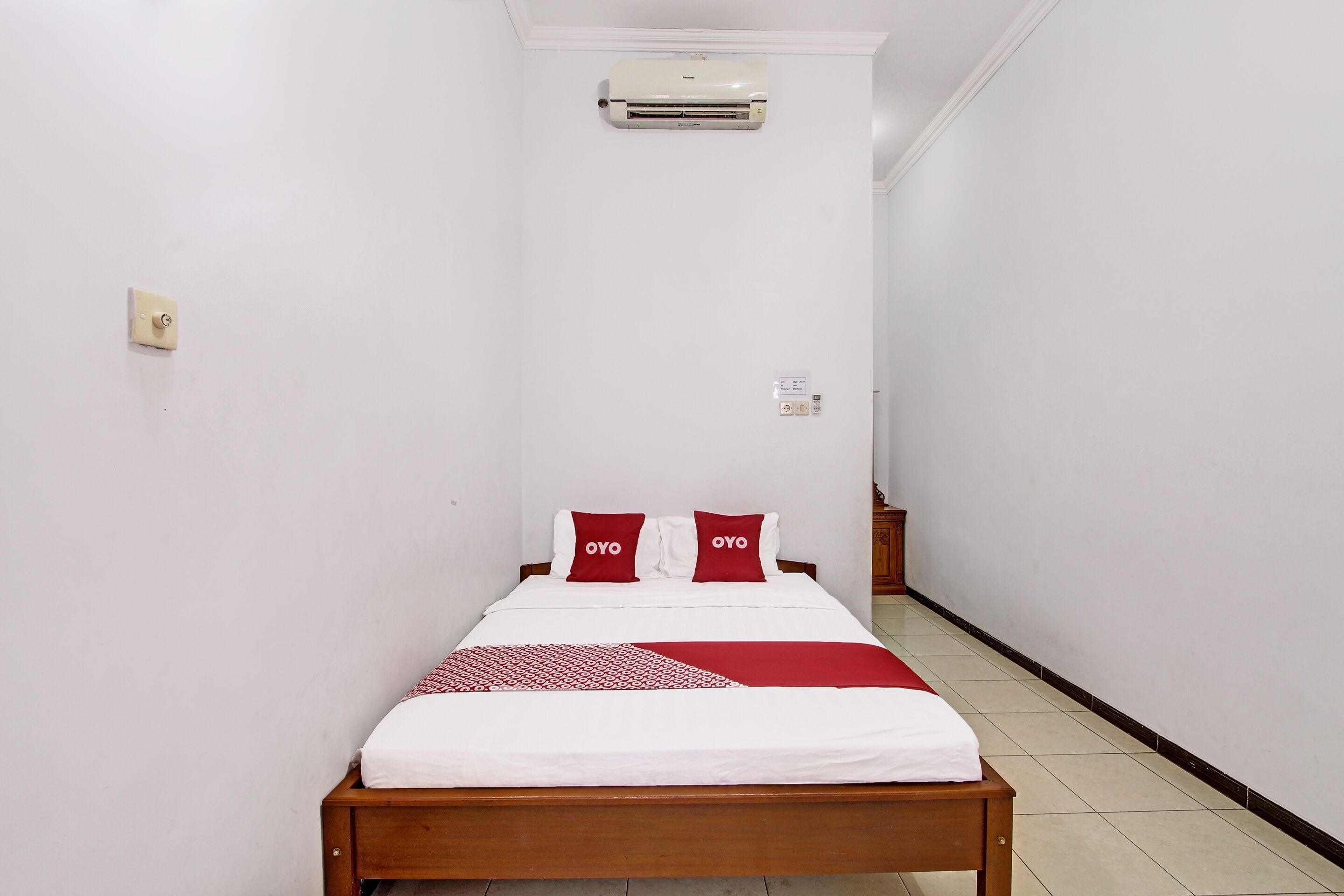 A narrow hotel room with a single bed, white walls, and a red and white bedspread.