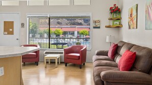 Ein modernes Wohnzimmer mit einem braunen Ledersofa, roten Sessel, einem kleinen weißen Tisch und einem großen Fenster mit Blick auf ein Gebäude und Bäume.