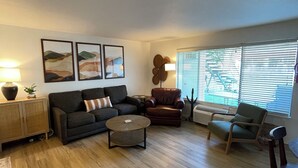 A living room with a sofa, coffee table, and two chairs. There are framed pictures on the wall, a lamp, and a radiator.