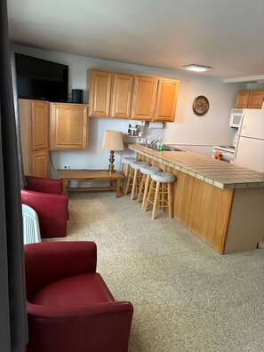 A compact kitchen with wooden cabinets, a coffee maker, a microwave, and a refrigerator. There are stools at the counter and a bench.
