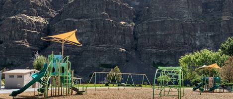 A playground with climbing structures and slides, set against a backdrop of rocky cliffs.