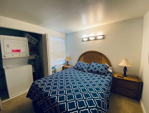 A bedroom with a bed, a nightstand, a lamp, a dresser, and a washing machine.