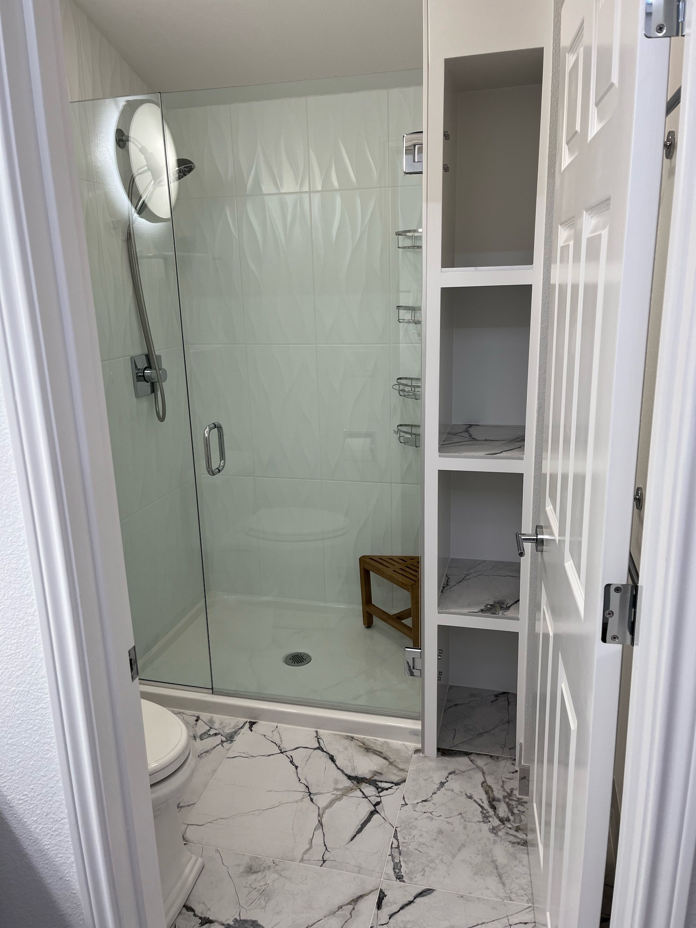 Une salle de bain avec une douche à parois de verre, des toilettes carrelées de marbre et un tabouret en bois.