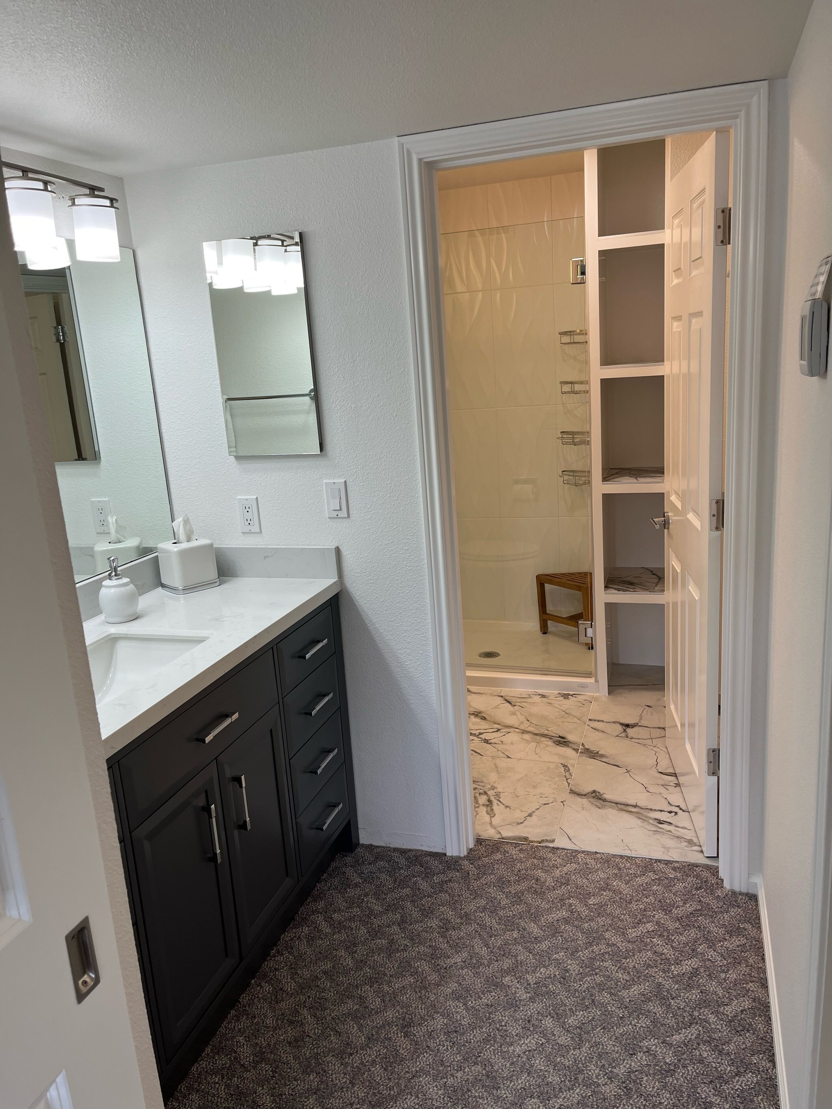 A bathroom with a white sink, dark cabinets, a large mirror, and a shower area visible through an open door.