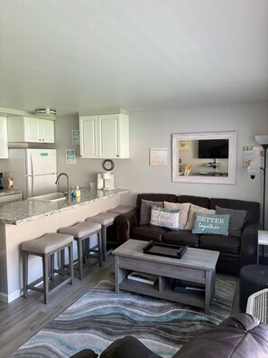A modern kitchen with white cabinets, a built-in sink, and a countertop. There is a dining area with a brown leather couch and a coffee table.