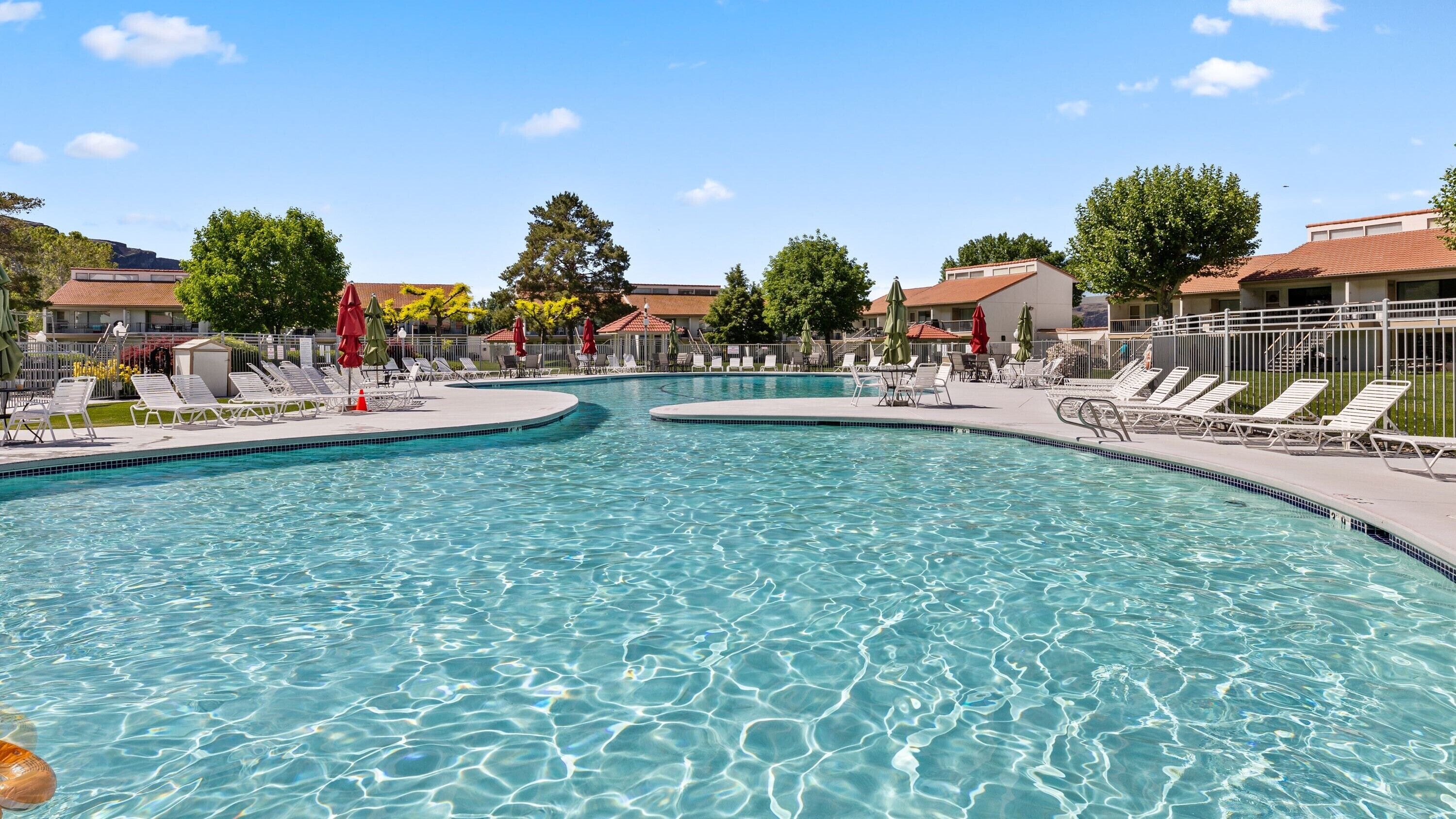 A clear swimming pool with lounge chairs and umbrellas, surrounded by buildings with red-tiled roofs.